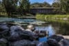 A section of the Milwaukee River courses through a park in Cedarburg, Wisconsin. In the foreground are small boulders and in the background a wooden bridge.