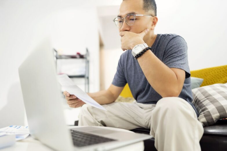 A man of east Asian ancestry wearing glasses sits in front of a laptop while examining a piece of paper.