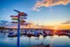 A pole with numerous signs pointing to various cities in the Midwest stands on a pier in Egg Harbor, Wisconsin, with boats docked in the background under a blue sky with a setting sun.