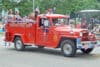Teenaged girls sit in the back of an old-fashioned bright red firetruck with the words "Trevor Fire Dept." written on the side. The car is decorated in 4th of July decorations and paradegoers are watching the scene from the sidewalks in the background.