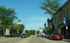 The main drag in downtown Sun Prairie, Wisconsin, with a row of two-story buildings on the right side, trees on both sides of the image, and a clear blue sky.