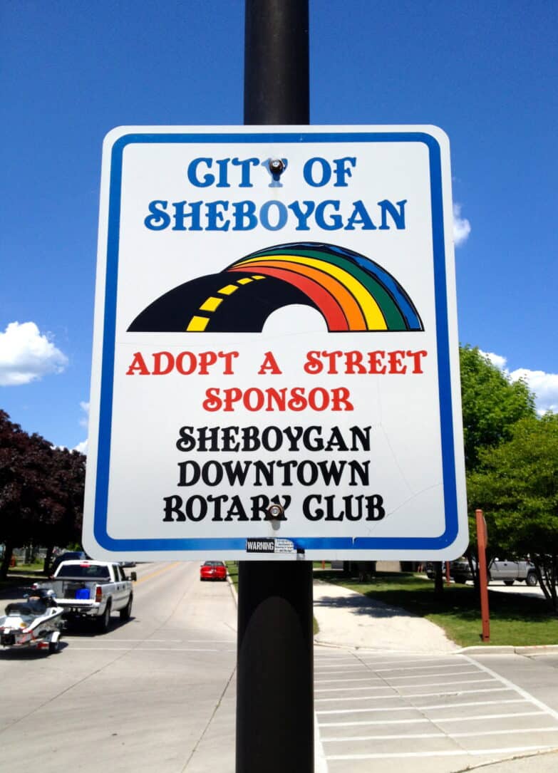 An outdoor sign that reads "City of Sheboygan. Adopt a Street Sponsor: Sheboygan Downtown Rotary Club. The sign has a white background with a rainbow and lettering in blue, red, and black.