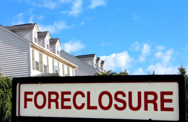 A sign in the foreground reads "Foreclosure" and behind it is a row of houses and a blue sky.