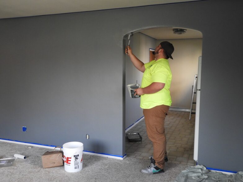 A man in a yellow T-shirt and tan pants painting the entrance to a room in a house.