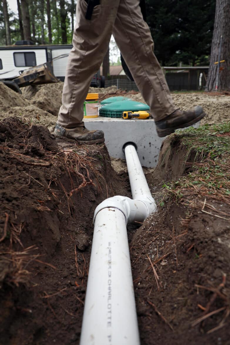 Inside of a ditch about one foot and two feet across deep runs PVC piping that connects to a septic tank, which is also below ground. A person is stepping over the ditch.
