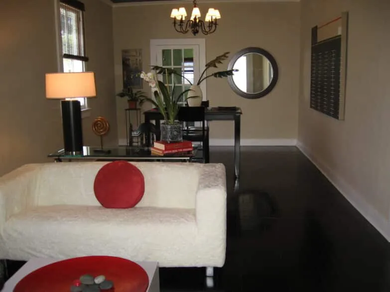 An immaculately staged dinning room in a house with ha white couch in the foreground, a mirror on the wall, a plant and dining room table.