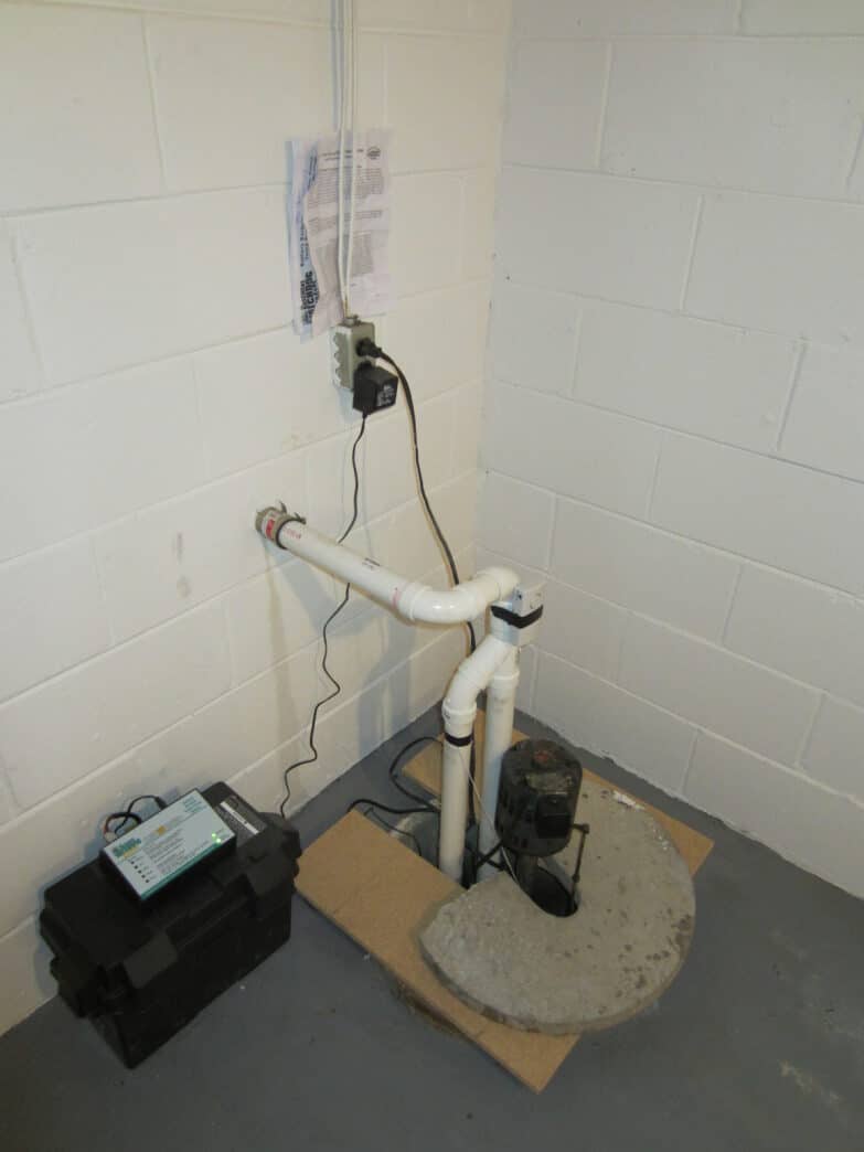 A sump pump in the corner of a clean basement with white brick walls.
