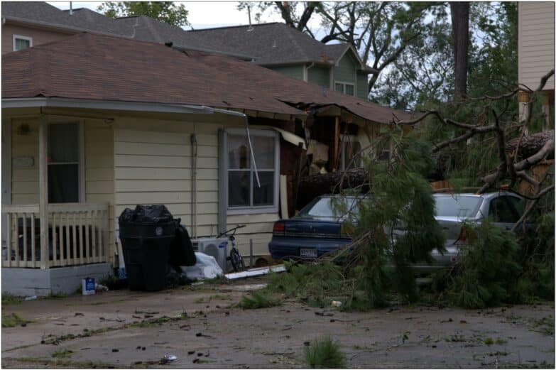 A fall tree lies near a house that it fell on, causing damage to the house.
