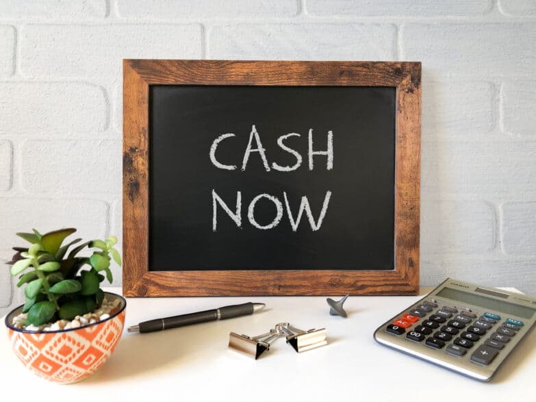 A small blackboard with "Cash Now" written on it in white chalk with a pen and calculator nearby on a table.