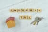 A photo of the words "property tax" made from scrabble pieces with a tiny fake house and keys flanking each side.