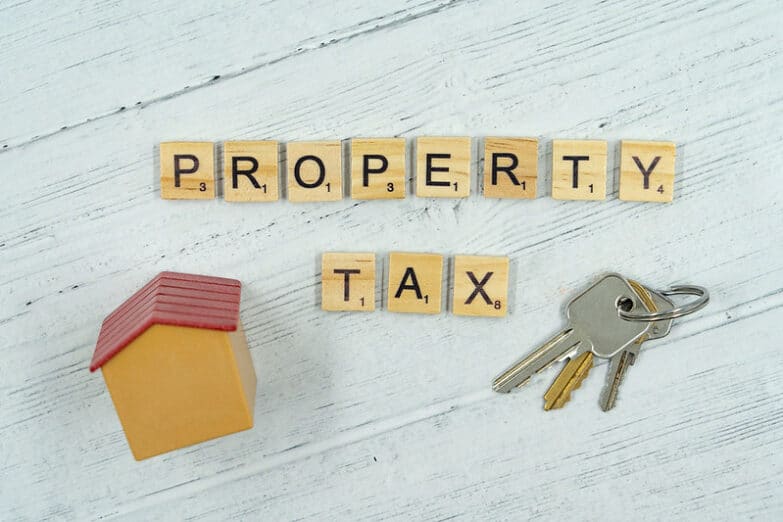 A photo of the words "property tax" made from scrabble pieces with a tiny fake house and keys flanking each side.