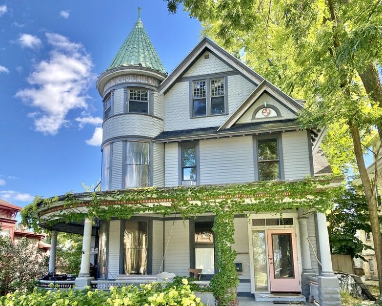 A photo of a 3-story Victorian house with vines and a wrap-around porch with a green tree off to the side.