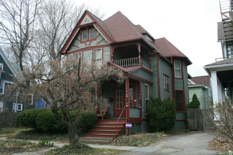A three-story house with a brown roof, a porch and a tree in front on a grey winter day.