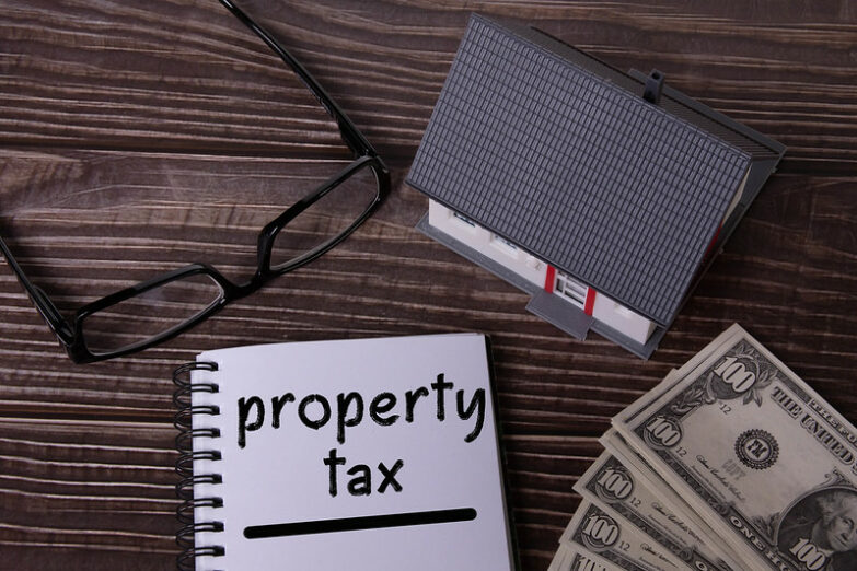 A notebook with the words "Property tax" written on it. Next to it is a little model house, $100 bills and a pair of glasses.